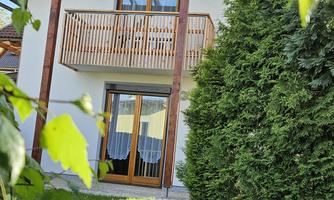 A modern house with wooden cladding and a balcony. The garden is green and well-maintained, surrounded by trees.