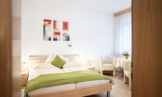 A bright room with a double bed and a green blanket. Next to the bed, there is a seating area with a table and plants.