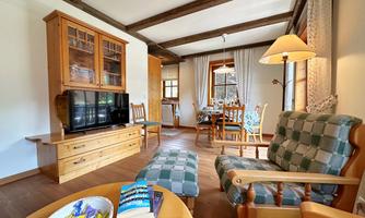 A cozy living room with wooden decor and a television. There are comfortable armchairs and a table with chairs in the background.