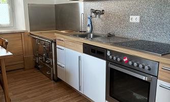 A modern kitchen with wooden cabinets and a silver countertop. It includes a stove, an oven, and is well-lit.