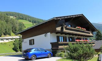 A beautiful house in the mountains with a blue car in front. The surroundings are green and the sun is shining.