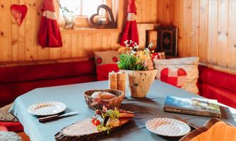 A cozy dining area in a wooden room with red curtains. The table is nicely set with plates and a floral decoration.