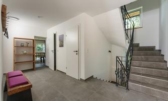 A bright entrance hall with a gray floor and bright lights. On the left, there is a shelf, and on the right, a staircase leads upstairs.