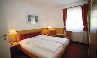 A cozy hotel room with a double bed and soft color tones. There are windows with curtains and a small table with chairs.