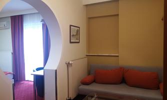 A spacious room with an orange sofa and a table. The room has large windows with curtains and a bright wall design.