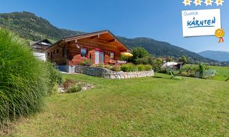A cozy wooden house in a green landscape. In the background, mountains and a blue sky can be seen.