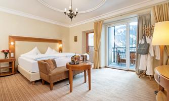 A cozy hotel room with a large bed, a table, and chairs. Large windows offer a view of the balcony and daylight floods in.