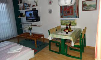 A cozy living space with a dining table, chairs, and a TV corner. On the table, there are wine bottles and a flower vase.