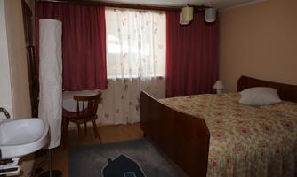 A cozy bedroom with a large bed and blooming curtains. Next to the bed, there is a chair and a nightstand with a lamp.