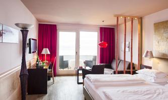 A modern hotel room with a double bed, elegant furnishings, and large windows. The furniture includes a desk, seating, and stylish decoration.