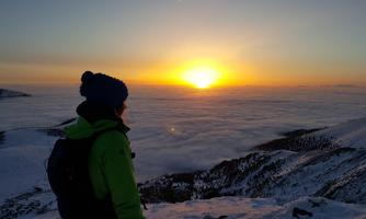 A person looks at a stunning sunset above the clouds. The landscape is covered in snow and the colors of the sky are warm and inviting.