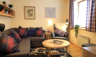 A cozy living room with a gray sofa and colorful cushions. There is a round table in the middle and windows with checkered curtains.