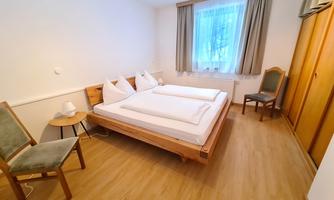 A simple, bright bedroom with two beds and a small table. The room has a large window with curtains and a wardrobe.