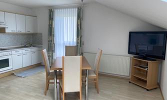 A bright apartment with a modern kitchen and dining area. There is a television in the room and large windows provide plenty of light.
