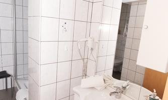 A modern bathroom with white tiles and a sink. On the left, there is a shower cabin and a mirror above the sink.