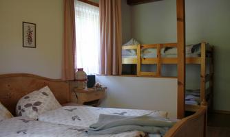 A cozy bedroom with a double bed and colorful pillows. In the background, there is a wooden bunk bed.
