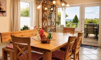 A cozy dining area with a large wooden table and chairs.  
A lot of light flows through the windows, and a beautiful view of nature is visible.
