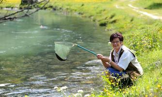 A person is sitting by the shore of a clear river, holding a net. Surrounded by colorful flowers and green grass, they are enjoying nature.