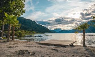 A picturesque lake with gentle hills in the background. Some palm trees line the shore, and a wooden dock invites you to relax.