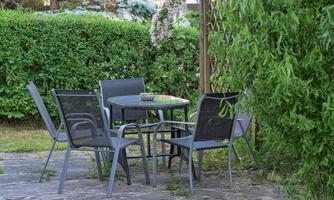 A round table with four chairs stands on a paved floor. In the background, green bushes and plants can be seen.