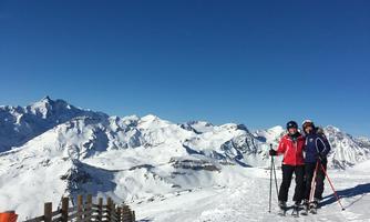 Two skiers are standing on a snow-covered slope. In the background, majestic mountains and a clear blue sky can be seen.