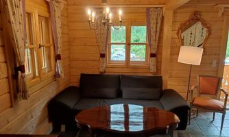 A cozy living area in a wooden cabin with a sofa and a wooden table. Soft light from a chandelier and large windows creates a warm atmosphere.
