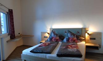 A cozy bedroom with a large double bed and colorful cushions. There are windows with curtains and a simple wooden floor.