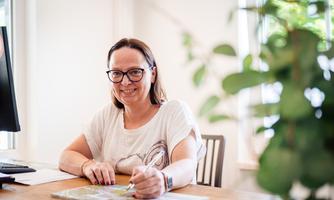 A smiling woman is sitting at a table and looking at the camera. Some documents are lying on the table and a plant is visible in the background.