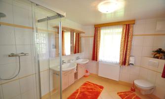 A modern bathroom with a shower, two sinks, and bright natural light. The orange floor mats add a warm accent.
