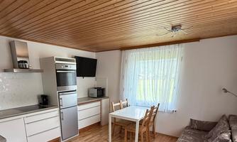 A modern kitchen with a dining table and chairs. The room has a cozy sofa and large windows that let in a lot of light.