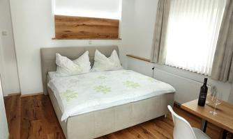 A modern bedroom with a large bed and fresh bed linen. Next to the bed is a table with a wine glass and a bottle of wine.