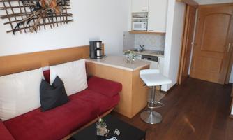A cozy living space with a red sofa and a small kitchenette. Next to the sofa is a modern bar stool and a small table.