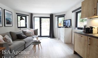 A modern living and dining room with a comfortable sofa and large windows. A television and a small kitchenette are also visible.