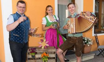 Three people are standing in front of an orange house. They are wearing traditional costumes and holding drinks as well as an accordion in their hands.