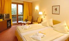 A cozy hotel room with two single beds and yellow curtains. There is a television, two chairs, and a balcony with a view outside.