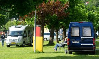 A campsite with motorhomes on a green lawn. In the background, there are trees and a relaxed person is sitting in a folding chair.