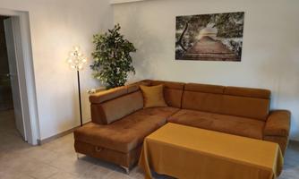 A cozy living room corner with a brown sofa and a colorful table. A picture of a forest path hangs on the wall.