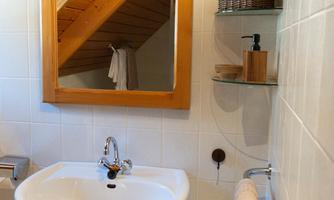 A modern bathroom with a sink, mirror, and wooden shelf. The walls are tiled in light colors, and a wooden ceiling gives the room a warm atmosphere.
