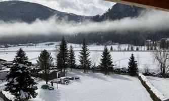 A snowy landscape with tall fir trees and mountains in the background. The sky is partially cloudy and there is a calm atmosphere in the air.