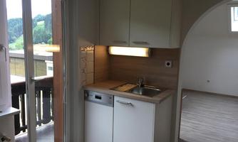 A modern kitchen with white cabinets and a sink. In the background, there is a light-filled room with access to the balcony.