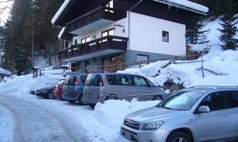 A snow-safe house in the mountains with several parked cars. The surroundings are surrounded by trees and snow.