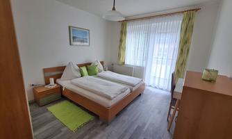 A modern bedroom with a double bed and two nightstands. Large windows with curtains let in plenty of daylight.
