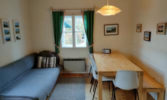 A cozy living space with a gray sofa and a dining table. The windows let in a lot of light and the walls are decorated with pictures.