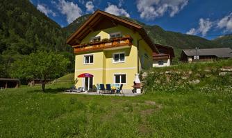 A beautiful, yellow house with a balcony and a terrace. It stands in a green landscape with mountains in the background.