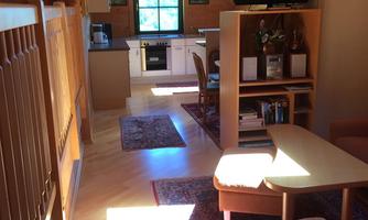 A bright and cozy room with wooden walls and a window that lets in light. In the foreground, furniture and a television are visible, while the kitchen is visible in the background.