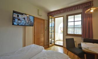 A bright room with a double bed, a television, and a window to the balcony terrace. There is a small table with chairs and plenty of natural light.