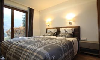 A cozy bedroom with a large double bed and a modern, minimalist design. The room has two windows with a view of nature and soft lighting.