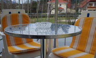 A small table with a glass ashtray and two chairs with orange-striped cushions is located on a balcony. In the background, houses and trees can be seen.