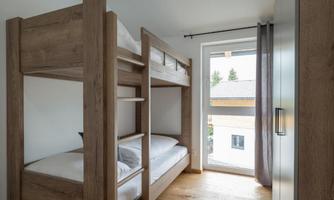 A cozy children's room with a wooden loft bed and two beds. Natural light enters the room through the window.