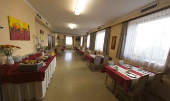 A spacious dining area with long tables, decorated with red tablecloths. Various dishes are displayed on the buffet, and large windows let in plenty of light.